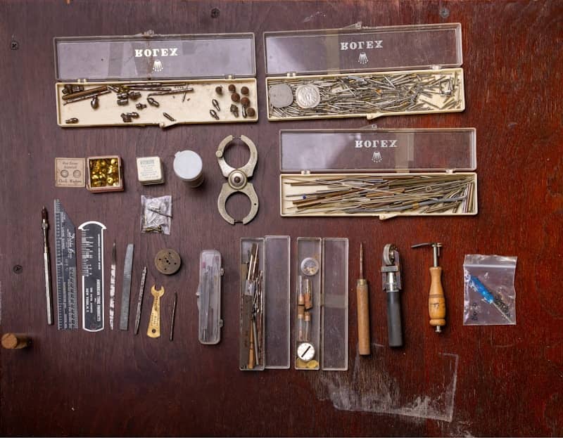 An assortment of watchmaker tools and small parts in plastic cases on a wooden table