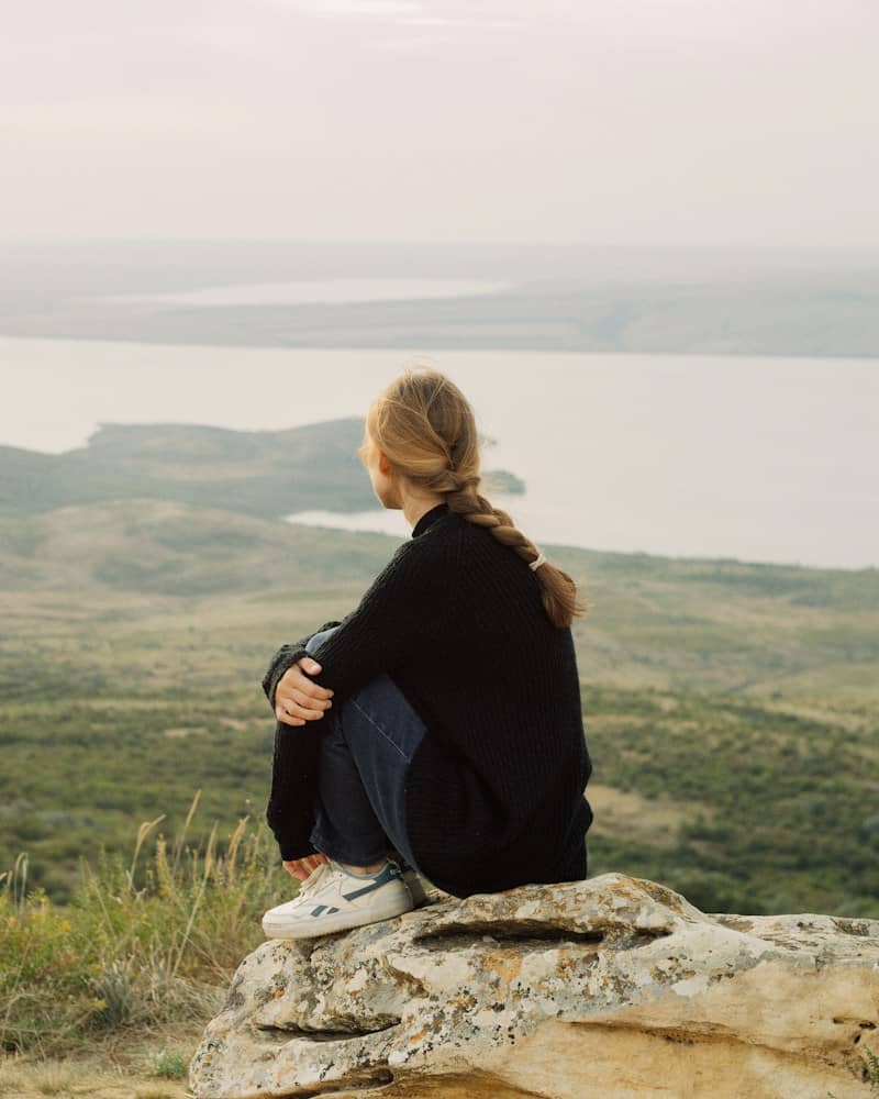 A person with a braided hairstyle sitting on a rock overlooking a lake