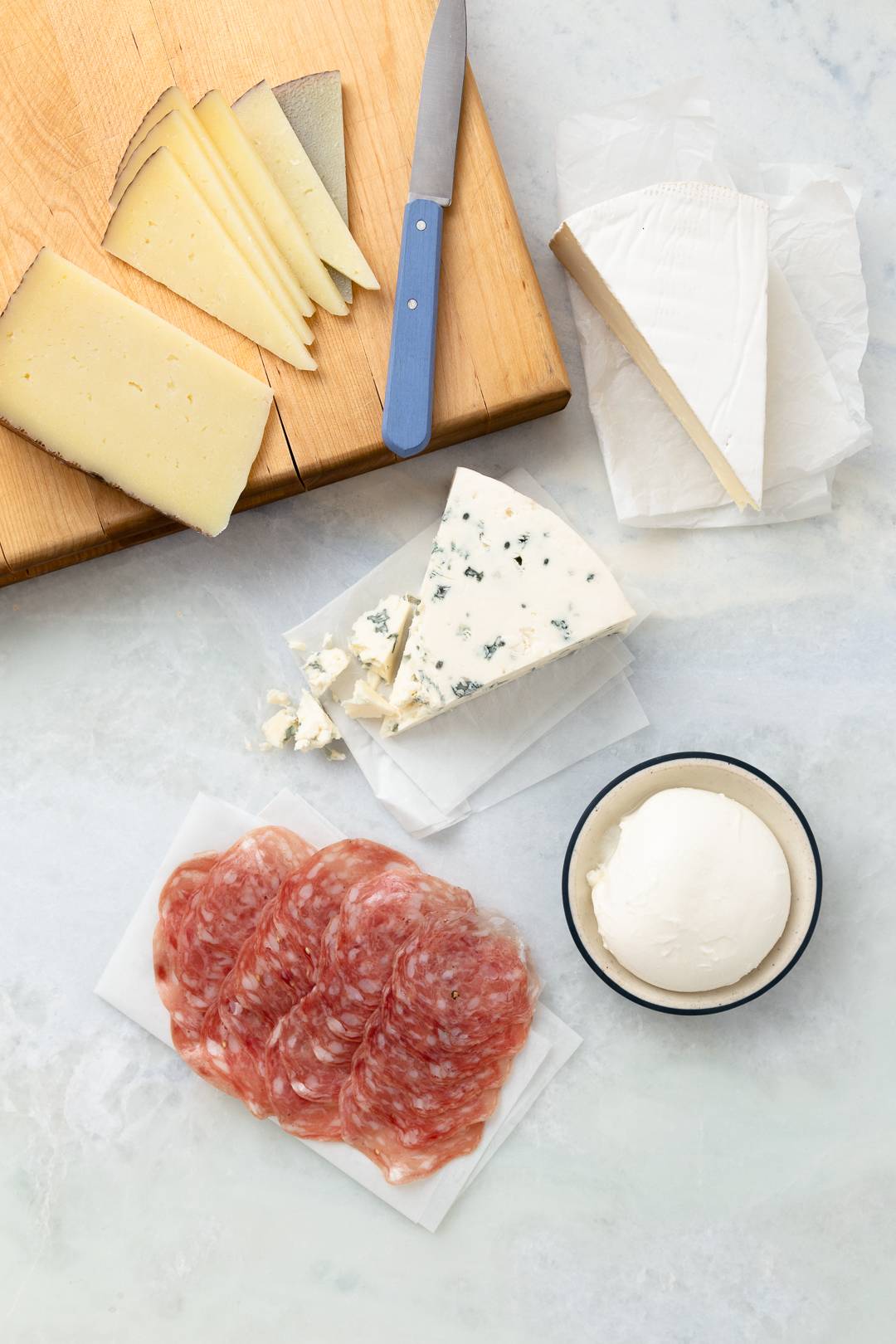 Overhead image of 4 wedges of different cheese, a ball of burrata, and a thinly sliced cured meat. 