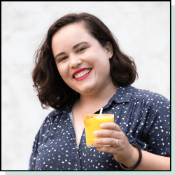 A woman with brown hair smiling and holding a yellow cocktail.