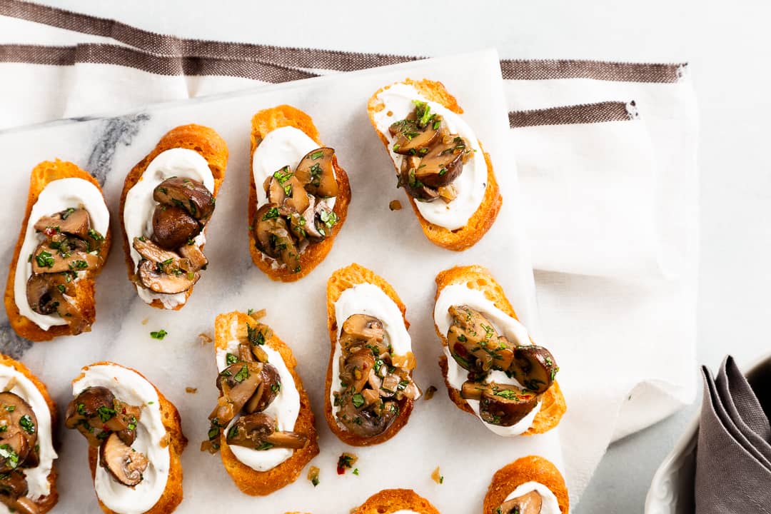 Multiple crostini on a marble slab with a creamy goat cheese on them and a mushroom topping.