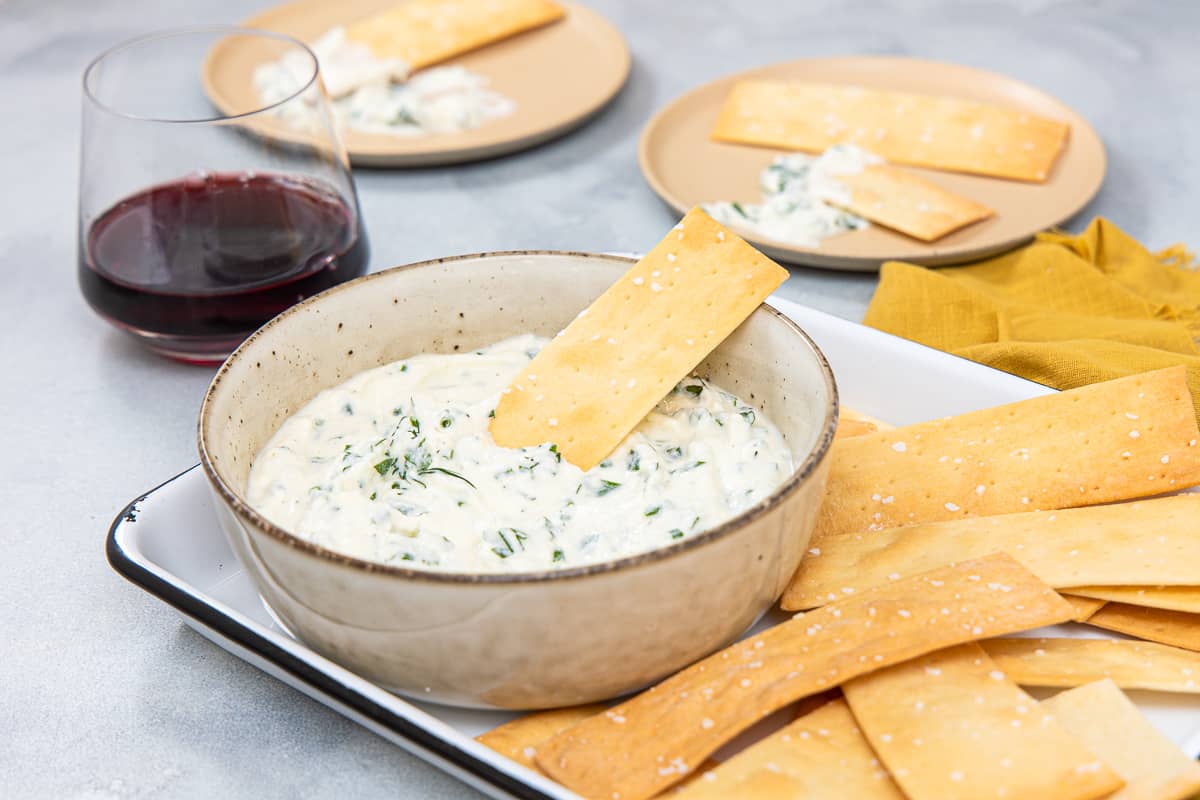 A bowl of a herby creamy dip with a cracker sticking out of it and other crackers surrounding it.