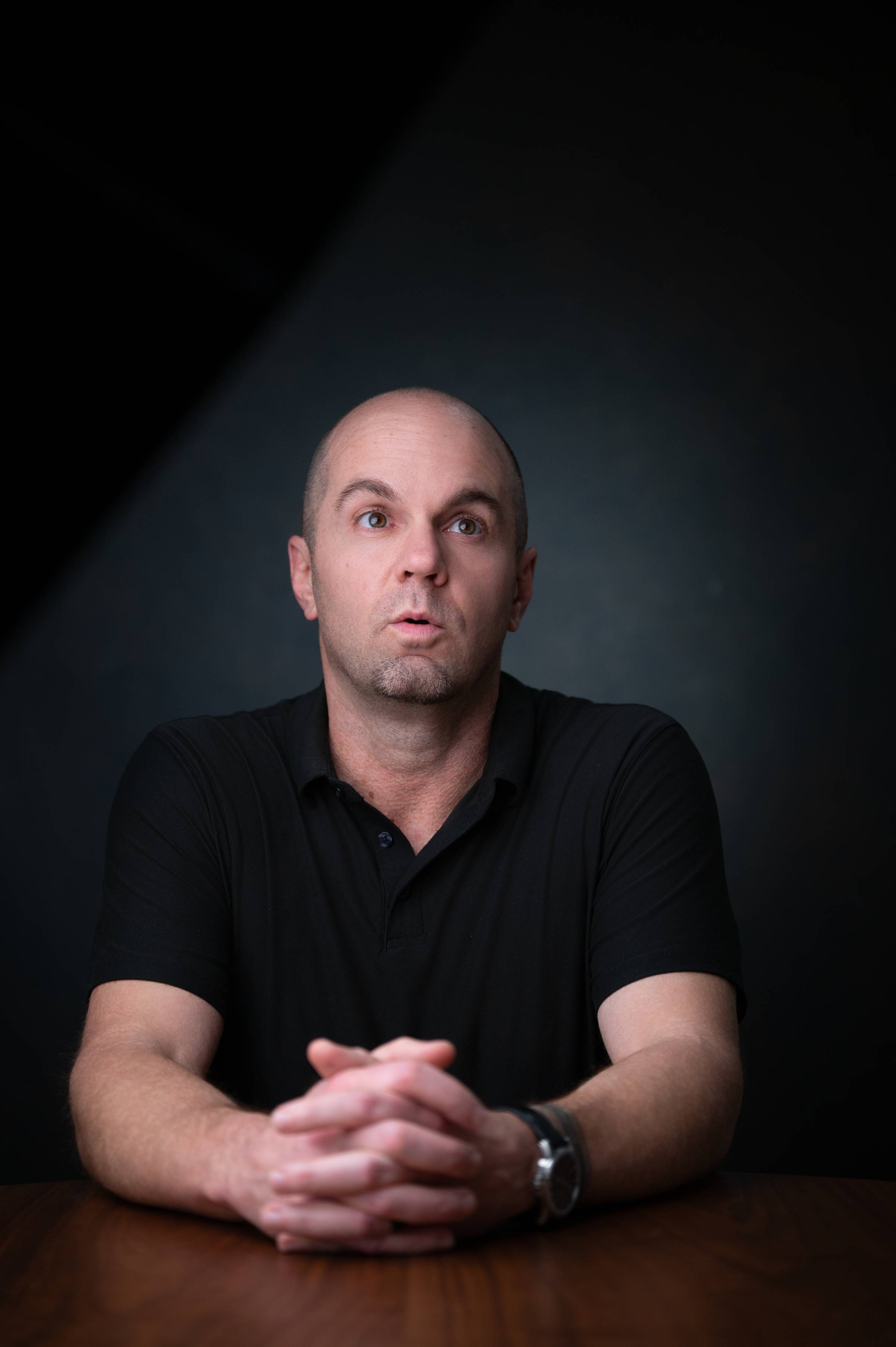 Entrepreneur and business advisor Nick Berry sits at a conference table with hands clasped, looking upward in a dark studio portrait setting.