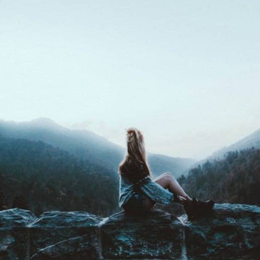 woman sitting on gray rock facing green mountains