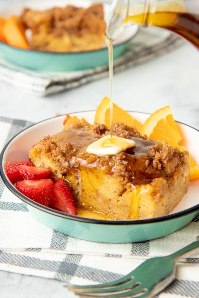Maple syrup pours over a serving of baked french toast topped with a pat of butter and plated with fresh strawberries and orange slices.